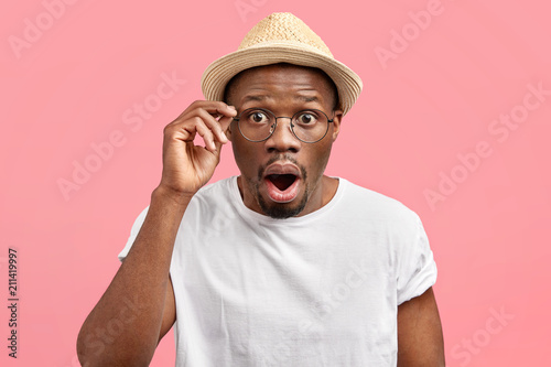 Fotografía Emotive stunned young African American male opens mouth and stares through glasses, being shocked by latest news, wears fashionable straw hat, stands alone against pink background