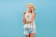 © oes - Young slender happy woman blond in shorts, a T-shirt and a straw hat carelessly stands on a blue isolated background and smiles. The model holds sunglasses near the face.