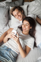 Happy young couple smiling lying in bed, checking email or social media relaxing on lazy morning, boyfriend and girlfriend resting at home reading news on smartphone, wife showing photos to spouse