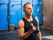 Handsome blond young man training biceps lifting dumbbells, standing in a gym, looking to a side