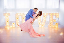 Groom gently tilting bride, holding her in his arms on background of large inscription love