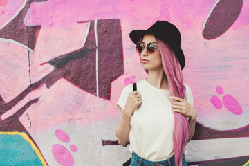 Beautiful stylish young hipster woman with long pink hair, hat and sunglasses on the street.