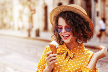Outdoor Close Up Portrait Of Young Beautiful Happy Woman Holding Ice Cream. Model Wearing Straw Hat, Heart Gradient Sunglasses, Polka Dot Yellow Dress, Posing In Street Of City. Copy Space For Text