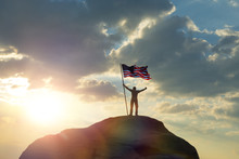 The Man Is Standing On The Top Of The Mountain, Holding His Hands Up With The Flag Of The US America Against The Sunset. Business Concept Idea, Success And Achievements, Career Ladder, Victory. 