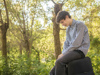 tired young teenage man sitting on large luggage case bag outdoor in the summer city street