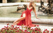 Fashion blonde woman in red maxi dress posing in garden.