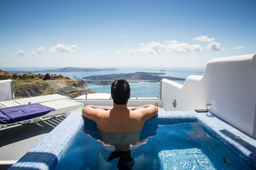 Dark haired man relaxing and contemplating beautiful scene from private pool in one of Oia, Santorini, Greece