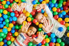 Above View Portrait Of Three Happy Little Kids In Ball Pit Smiling At Camera Raising Hands While Having Fun In Children Play Center, Copy Space