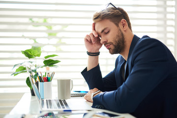 Wall Mural - Side view profile of exhausted businessman sitting at laptop during working day. He leaning at hand with his eyes closed feeling drained and sleepy