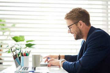 Wall Mural - Profile of smiling male sitting at desktop and touching notebook. There are laptop, coffee mug and colorful markers on table