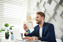Side View Profile Of Smiling Businessman Chatting On Phone In Office. He Is Sitting At Desktop With Laptop In Front Of Him And Typing On Screen With Joy