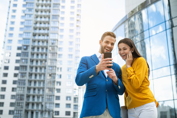 Let me show you my new device. Portrait of excited young man is expressing happiness while keeping smartphone. Girl is looking at technology with interest and laughing. Skyscraper on background 