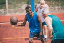 Multiracial Elderly Men Playing Basketball Together On Playground On Summer Day
