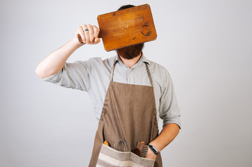 Young chef bearded man covers his face with a wooden cutting board. Man wearing a shirt and an apron on white background.