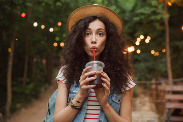 Poster - Photo of lovely summer woman 18-20 wearing straw hat, looking at you while drinking beverage from plastic cup during rest in green park with colorful lamps background