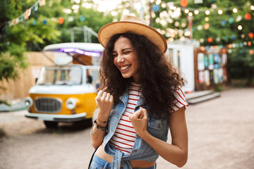 Poster - Photo of cheerful happy woman 18-20 with curly brown hair, clenching fists and expressing delight while walk through hipster spot or modern park with colorful lamps background