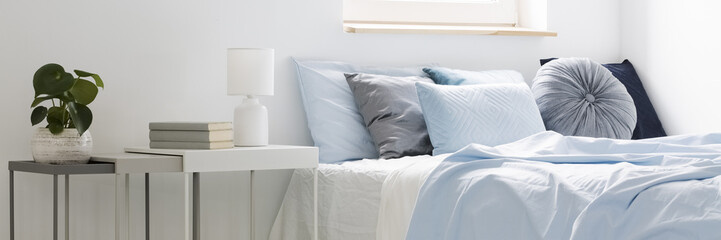 real photo of a bed with blue bedding and cushions standing next to white tables with books, lamp an