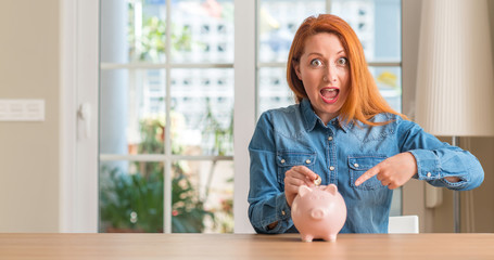 Sticker - Redhead woman saves money in piggy bank at home very happy pointing with hand and finger