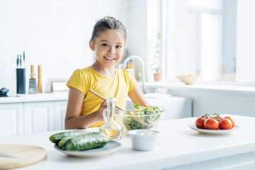 smiling little girl preparing salad at kitchen and looking at camera