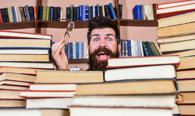 Leinwandbilder - Man on happy face between piles of books in library, bookshelves on background. Scientific research concept. Teacher or student with beard wears eyeglasses, sits at table with books, defocused.