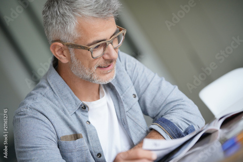 Cuadros en Lienzo Middle-aged guy with trendy eyeglasses reading magazine
