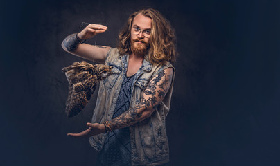 Wall Mural - Portrait of a tattoed redhead hipster male with long luxuriant hair and full beard dressed in a t-shirt and jacket holds a keeps the scarecrow of an owl in a studio. Isolated on the dark background.