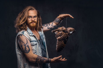 Wall Mural - Portrait of a tattoed redhead hipster male with long luxuriant hair and full beard dressed in a t-shirt and jacket holds a keeps the scarecrow of an owl in a studio. Isolated on the dark background.