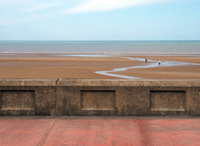 The Concrete Seawall In Blackpool Behind The Pedestrian Walkway With The Beach At Low Tide And Calm Summer Sea With Blue Sky And Clouds