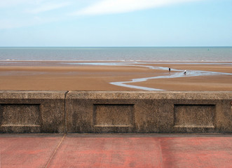 the concrete seawall in blackpool behind the pedestrian walkway with the beach at low tide and calm summer sea with blue sky and clouds