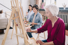 Portrait Of Art Students Sitting In Row And Painting At Easels In Art Studio, Focus On Smiling  Adult  Woman Enjoying Work In Center, Copy Space