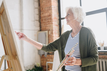 Waist up side view portrait of white haired senior woman holding palette painting pictures at easel in  art studio standing against windows in sunlight, copy space