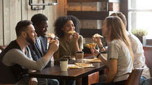 Smiling Multiracial Friends Eating Pizza And Drinking Coffee, Laughing And Having Fun In Restaurant, Diverse Millennial Colleagues Enjoying Lunch During Work Break Sitting At Coffee Table In Loft Cafe