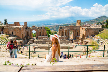 Wall Mural - Jule 04, 2018. Taormina, Italy. Beautiful girl in a long white dress and white summer hat sitting at the caldron of the ancient Greek Theater of Taormina in Italy.