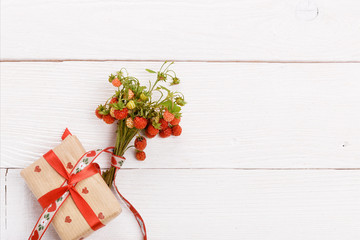 Wall Mural - Wild strawberries bouquet and gift box with red ribbon and heart on white wooden background. Overhead top view, flat lay. Copy space. Wedding, Birthday, Mother's, Valentines, Women's Day concept