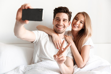 Poster - Cheerful loving couple lies in bed take a selfie showing peace gesture.