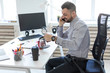 © Ivan Traimak - A young man in the office sits at a table, talking on the phone and pouring coffee into a cup.