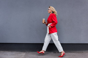 side view of attractive woman in red jacket walking with coffee in paper cup on street