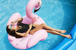 Hot and fashionable brunette, fitness model girl with perfect sexy body in stylish black bikini and glamorous sunglasses, tanning on an inflatable pink flamingo and posing at the swimming pool