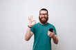 © Vulp - Portrait of a happy young bearded hipster man wearing eyeglassses and showing ok gesture and holding  phone. looking at camera isolated over white background