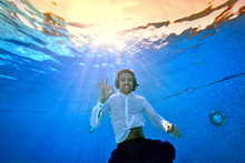 A Young Smiling Man With A Headset On His Head Swims And Poses Underwater In The Pool In A White Shirt Against The Yellow Sunset. He's Looking At The Camera. Portrait. Underwater Photography