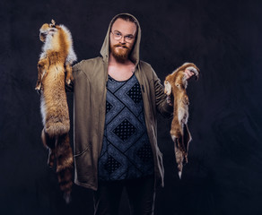 Wall Mural - Portrait of a redhead hipster guy with full beard in glasses dressed in hoodie and t-shirt standing with fox skins in his hands at a studio. Isolated on the dark background.