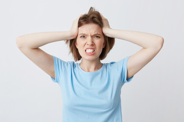 Wall Mural - Portrait of angry irritated young woman in blue tshirt standing with hands on head and feels crazy isolated over white background Looks directly in camera