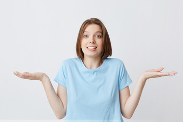 Wall Mural - Portrait of confused cute young woman in blue t shirt shrug her shoulders and hold copyspace on both palms isolated over white background