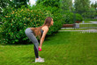 © Виталий Сова - A dark-haired woman coach in a sporty short top and gym leggings makes a deadlift with dumbbells   on a summer day in a park on a green lawn
