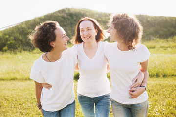 Family portrait of a positive mother hugs her daughters in white T-shirts and jeans against the backdrop of a green hill on a summer sunny day.