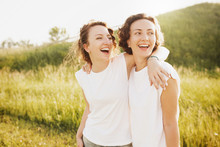 Portrait Of Cute Young Beautiful Curly-haired Girls Sisters Standing Into The Distance Dressed In A White T-shirt Posing Against A Green Hill