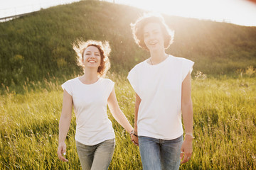 Beautiful young positive female sisters walking holding hands dressed in white t-shirts and jeans against the backdrop of a green scenic hill sunny summer day