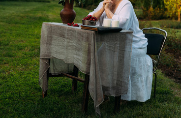 grandmother sitting in a chair at the table on which the cherry is lying, in the garden and old album, book. grandmother in a white vintage dress, summer, warm day. on the table there are two glasses