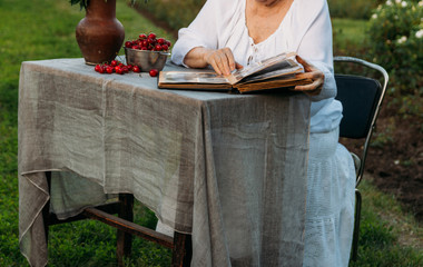 grandmother sitting in a chair at the table on which the cherry is lying, in the garden and watching an old album with photos, recounts and remembers the family's story. grandmother in a white vintage