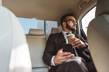 handsome businessman sitting in car with coffee in paper cup and smartphone